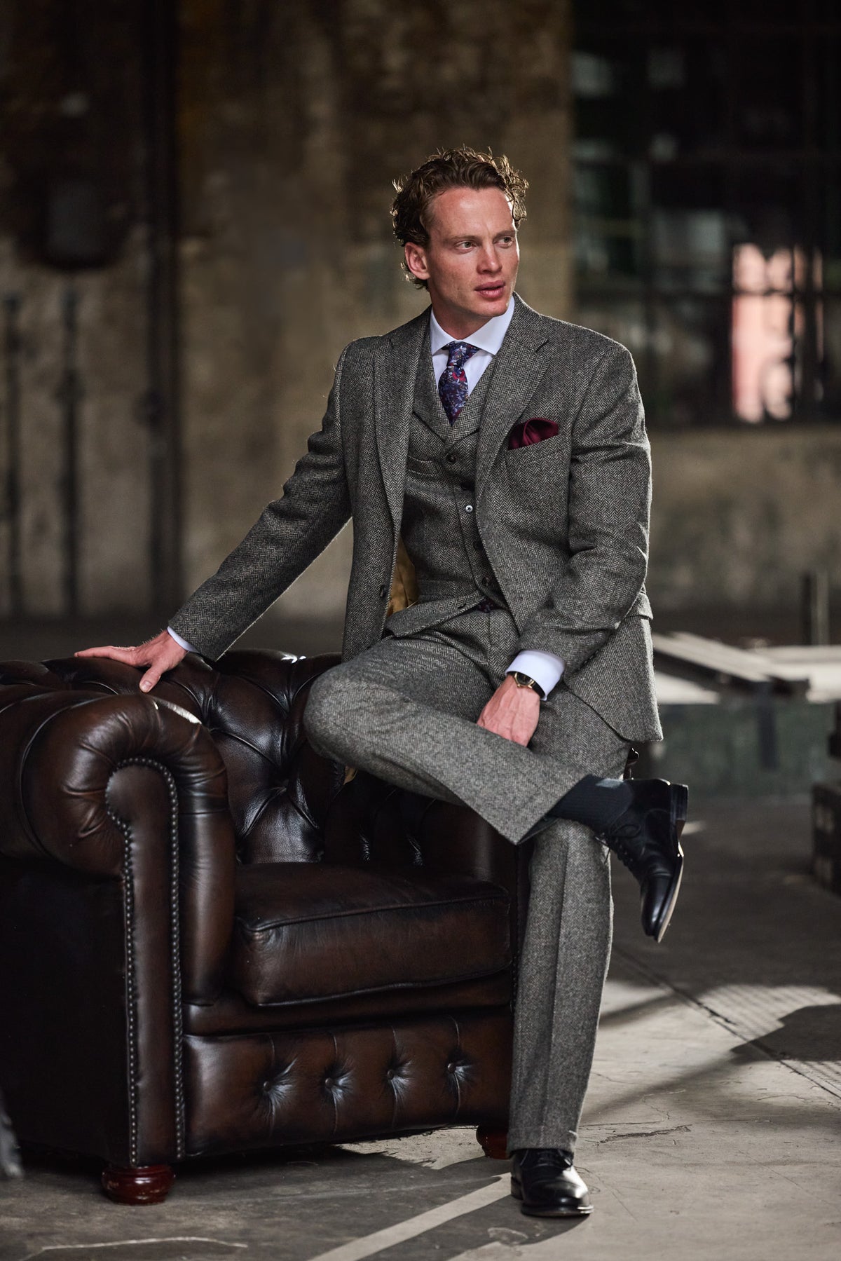 Man in a grey three-piece tweed suit with tie and pocket square by a leather armchair — John Crocket