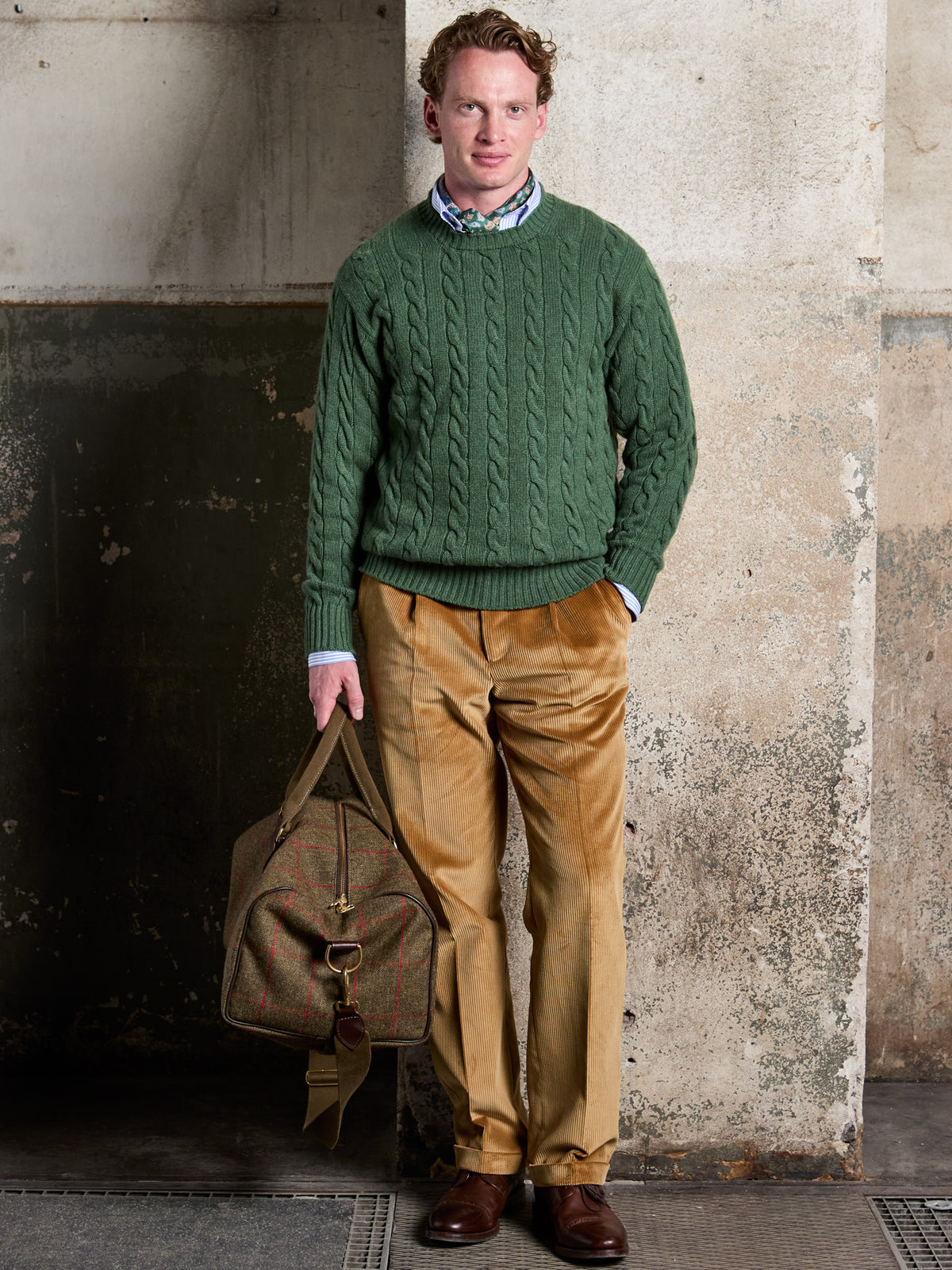 Weekender in Lovat Tweed, Olive, in the hand, combined with a green cable-knit jumper and corduroy trousers – John Crocket