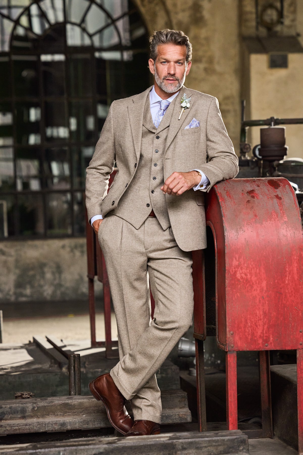 Man in a beige three-piece tweed suit with tie and buttonhole, leaning on a red machine — John Crocket