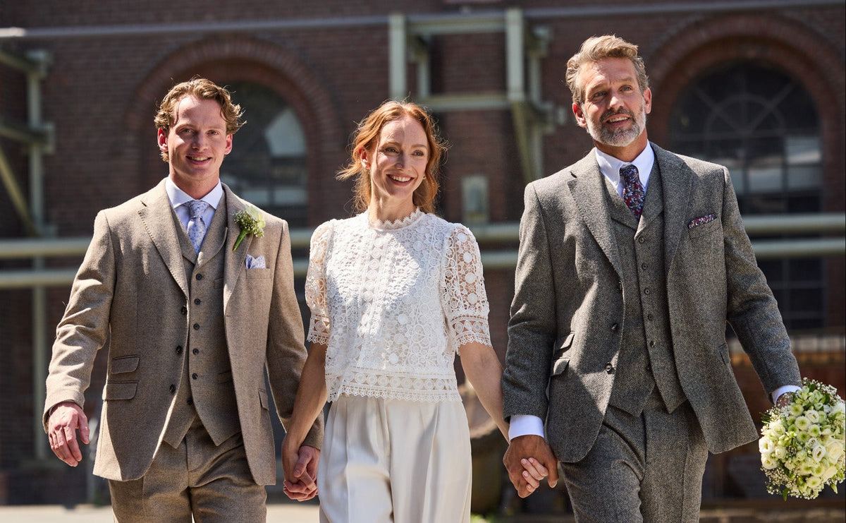 Woman in a white lace blouse between two men in three-piece tweed suits by a brick building — John Crocket