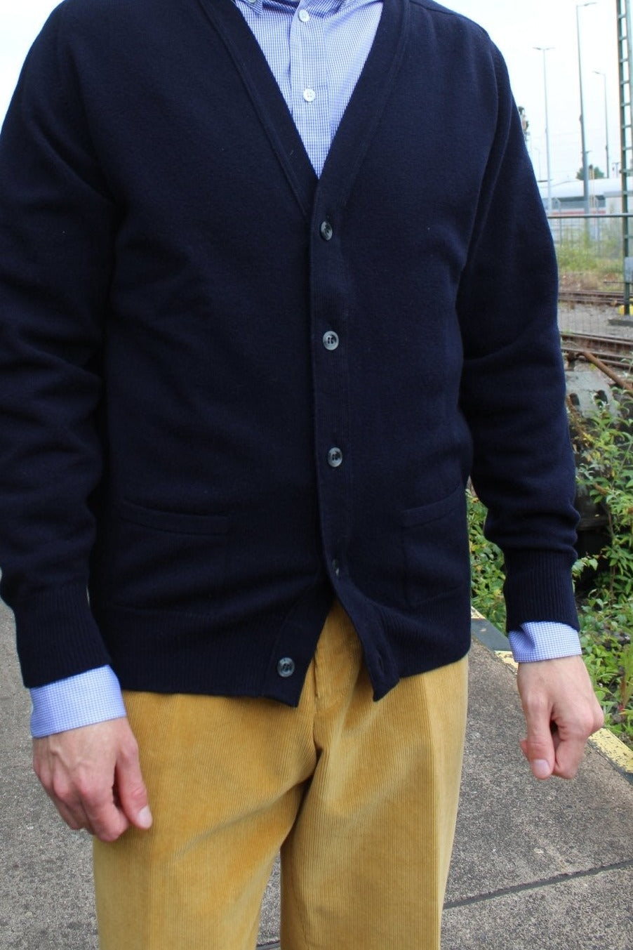 Lambswool cardigan in navy, worn by the model with a light blue shirt and yellow corduroy trousers – John Crocket