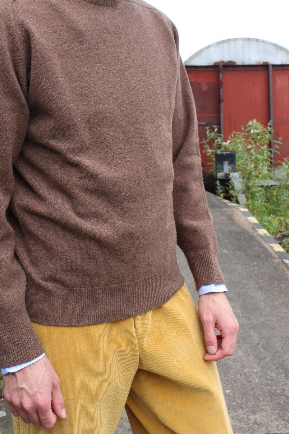 Lambswool crew-neck jumper in brown, worn by the model beside a brick wall with a light blue shirt and yellow corduroy trousers – John Crocket