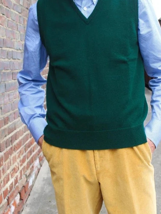 Lambswool spencer met V-hals in donkergroen, gedragen door model voor een bakstenen muur, met lichtblauw overhemd en gele ribbroek – John Crocket