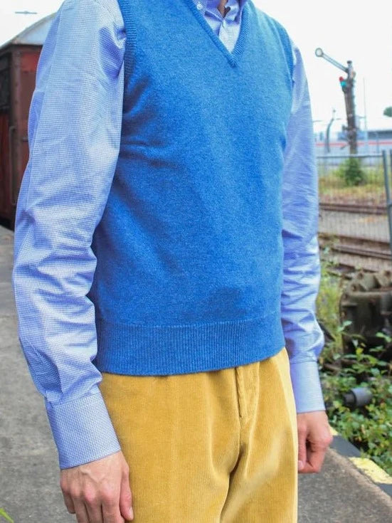 Lambswool spencer met V-hals in blauw, gedragen door model dat opzij kijkt, met lichtblauw overhemd en gele ribbroek – John Crocket