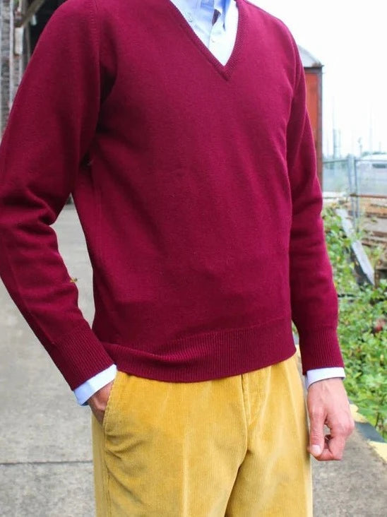 Lambswool V-neck jumper in burgundy, worn by the model with a light blue shirt and yellow corduroy trousers – John Crocket